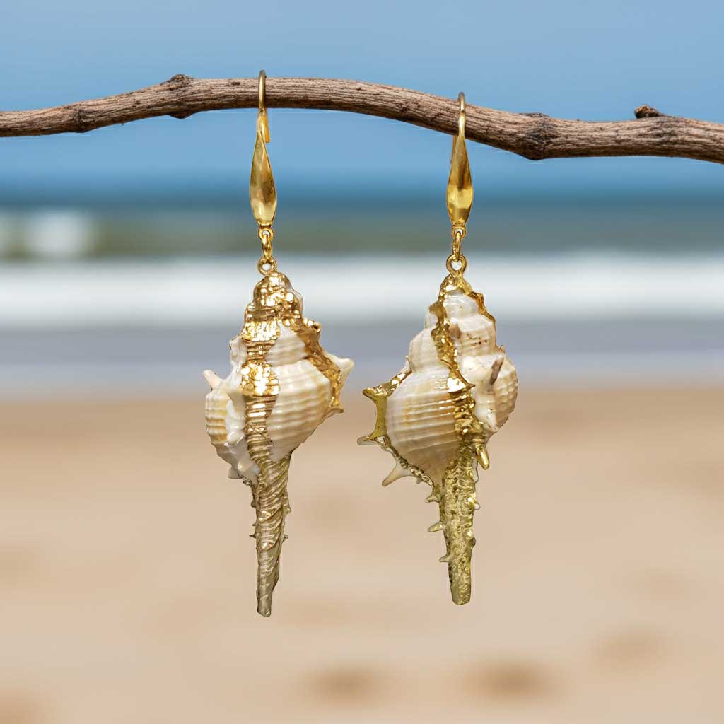 Lange schelpen oorbellen met witte schelp en goud edelstaal hangend aan takje met achtergrond zee| Shell Cabrit Gold