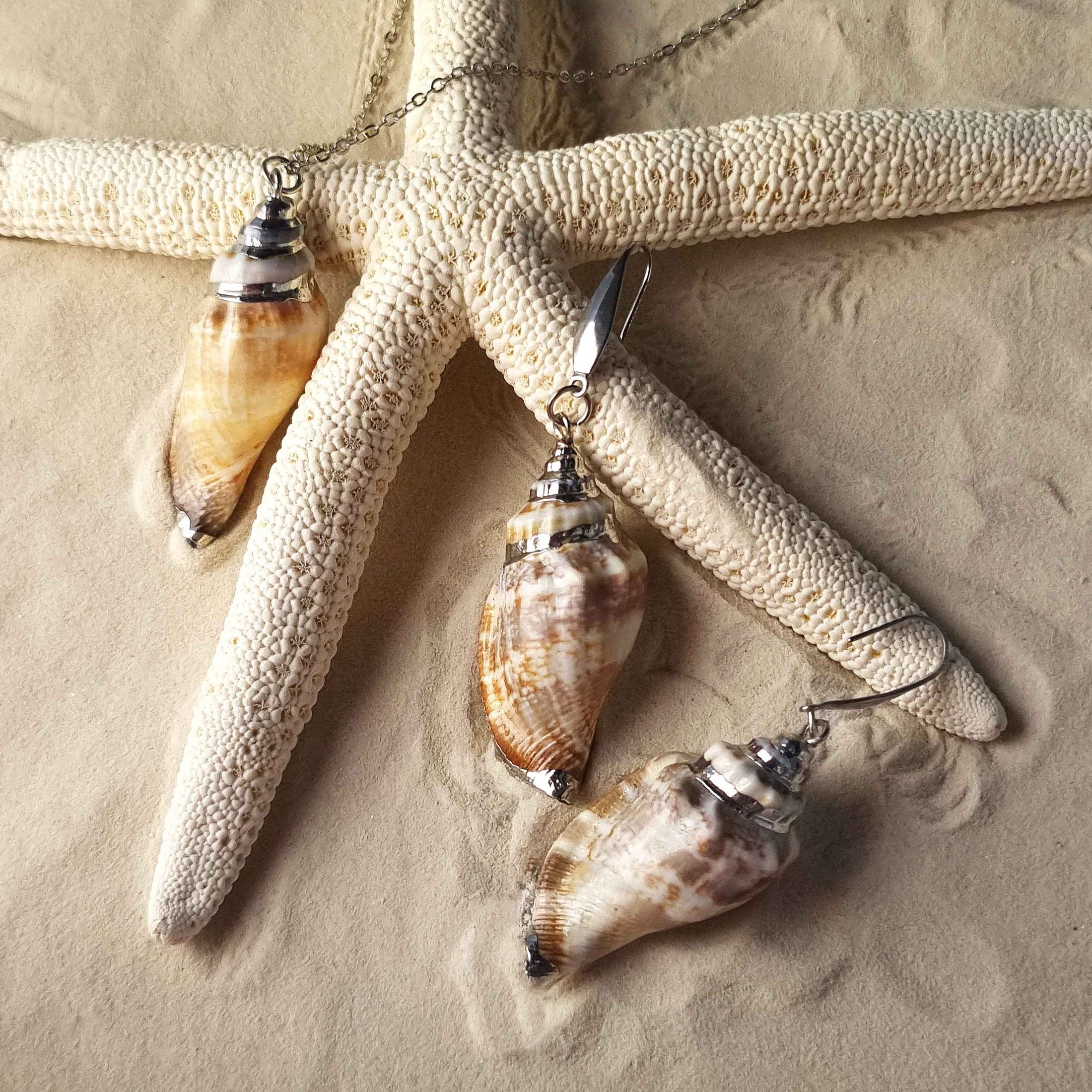Schelpen sieraden set bestaande uit een schelpen ketting en schelpen oorbellen met licht bruine schelp en zilver edelstaal liggend op zand | set Seashell Silver