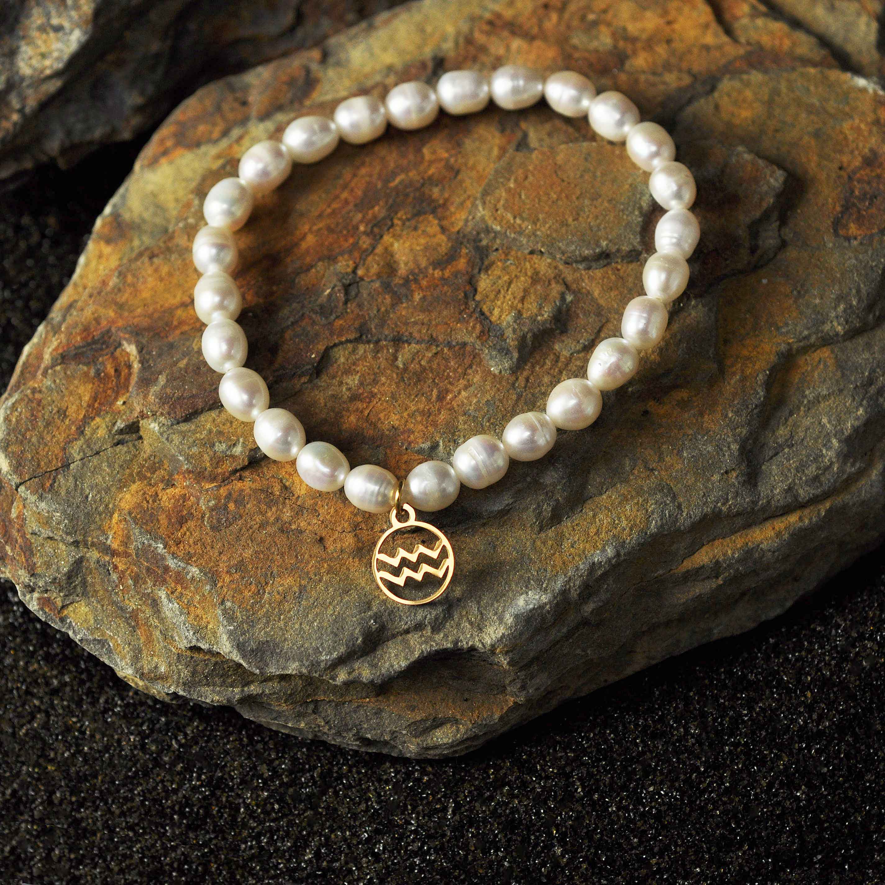 Wit zoetwater parel armband met goud edelstalen symbool van sterrenbeeld Waterman liggend op steen | Parel armband Gouden sterrenbeeld Waterman