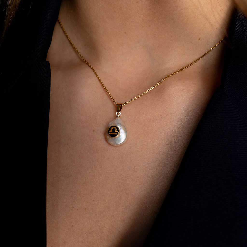 Detail van zoetwater parelketting met witte parel hanger met gouden symbool van sterrenbeeld weegschaal en goud edelstaal op model met colbertje | Gouden Sterrenbeeld Weegschaal