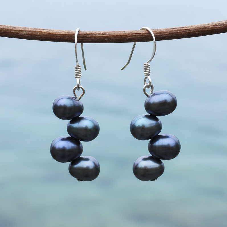 Zilveren fish hook oorbellen met vier donkere peacock parels aan takje boven lichtblauw water
