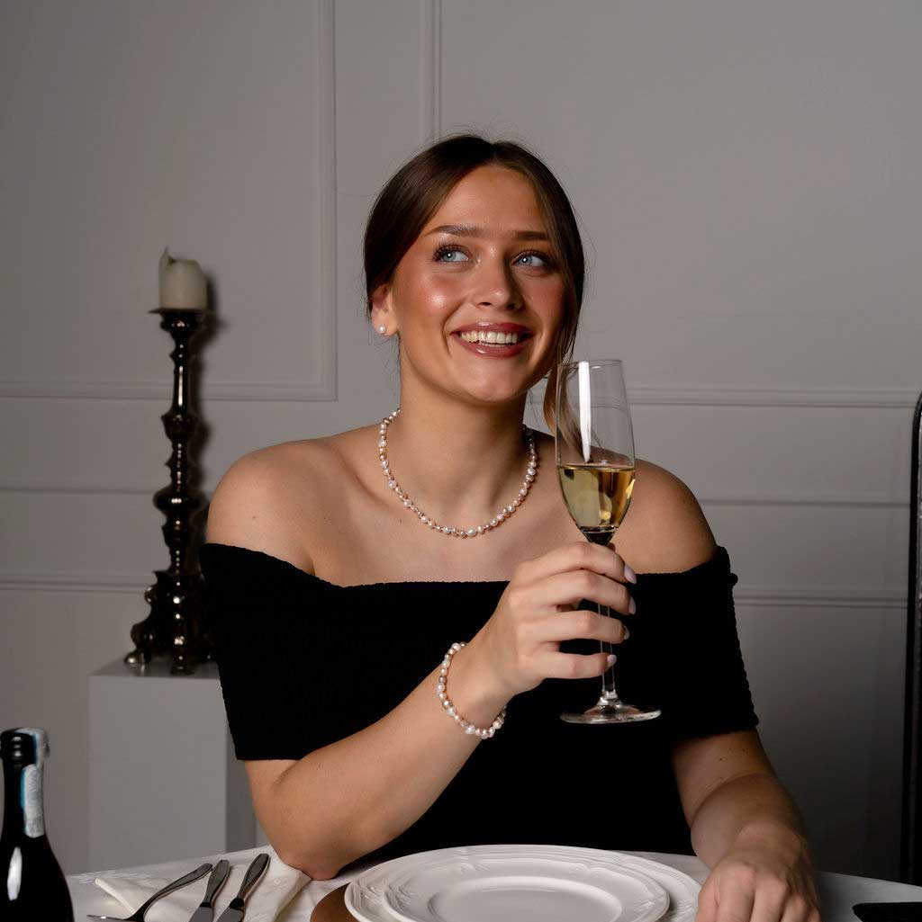 Handgeknoopte zoetwater parel set bestaande uit parelketting en parel armband met witte, zalm en roze parels en sterling zilver (925) op model aan tafel met glas champagne | set Pearl Colors Small