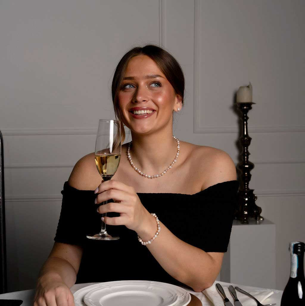 Handgeknoopte zoetwater parel set bestaande uit parelketting en parel armband met witte, zalm en roze parels en sterling zilver (925) op model aan tafel met glas champagne | set Pearl Colors Small