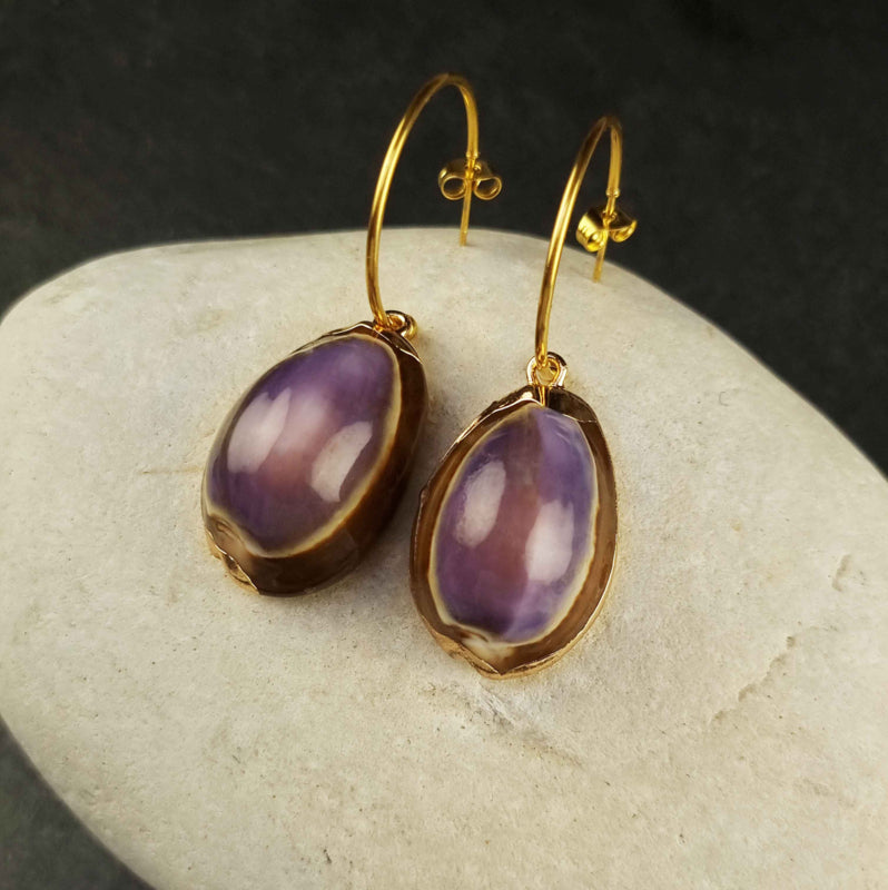 Gouden oorringen met paarse schelpen en goud edelstaal liggend op witte kei | Shell Cowrie Purple