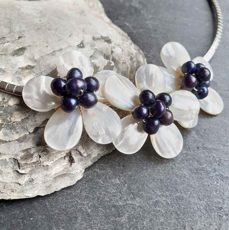 Detail van zoetwater parelketting met blauwe parels, wit parelmoer in de vorm van een bloem en zilver edelstaal liggend tegen schelp | Florien B