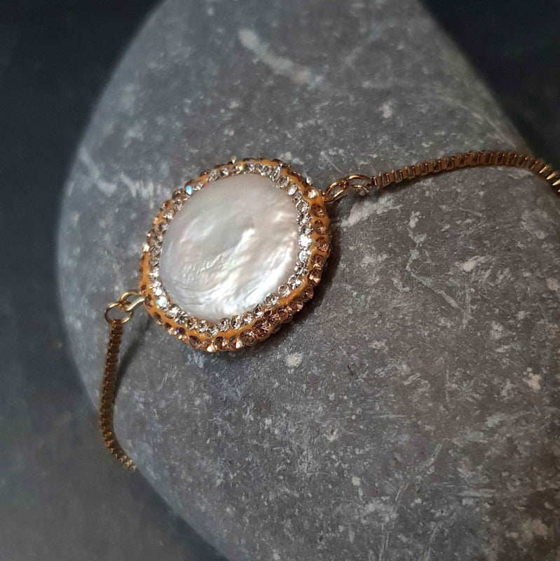 Wit zoetwater parel armband met goud edelstaal, stras steentjes en een schuifslotje liggend op kei | Bright Golden Pearl