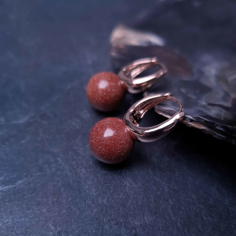 Bruine edelstenen oorbellen met goudvuur / goudsteen en rose goud liggend tegen schelprand | Rose Gold Goldstone Ball