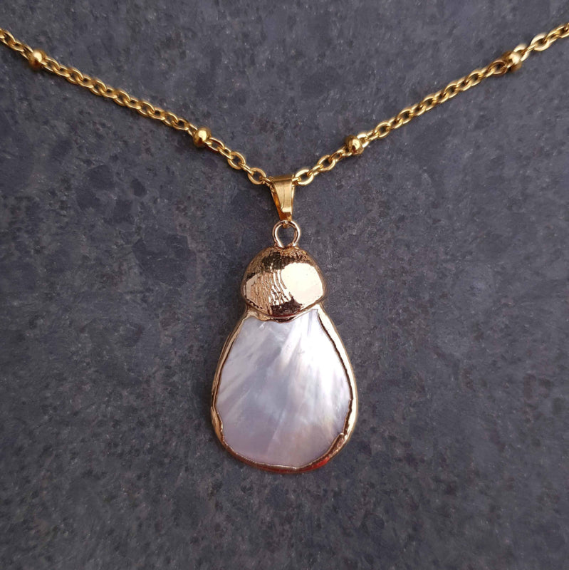 Detail van witte parelmoeren ketting met wit parelmoer hanger aan gouden edelstalen ketting liggend op leisteen | Gold Shell