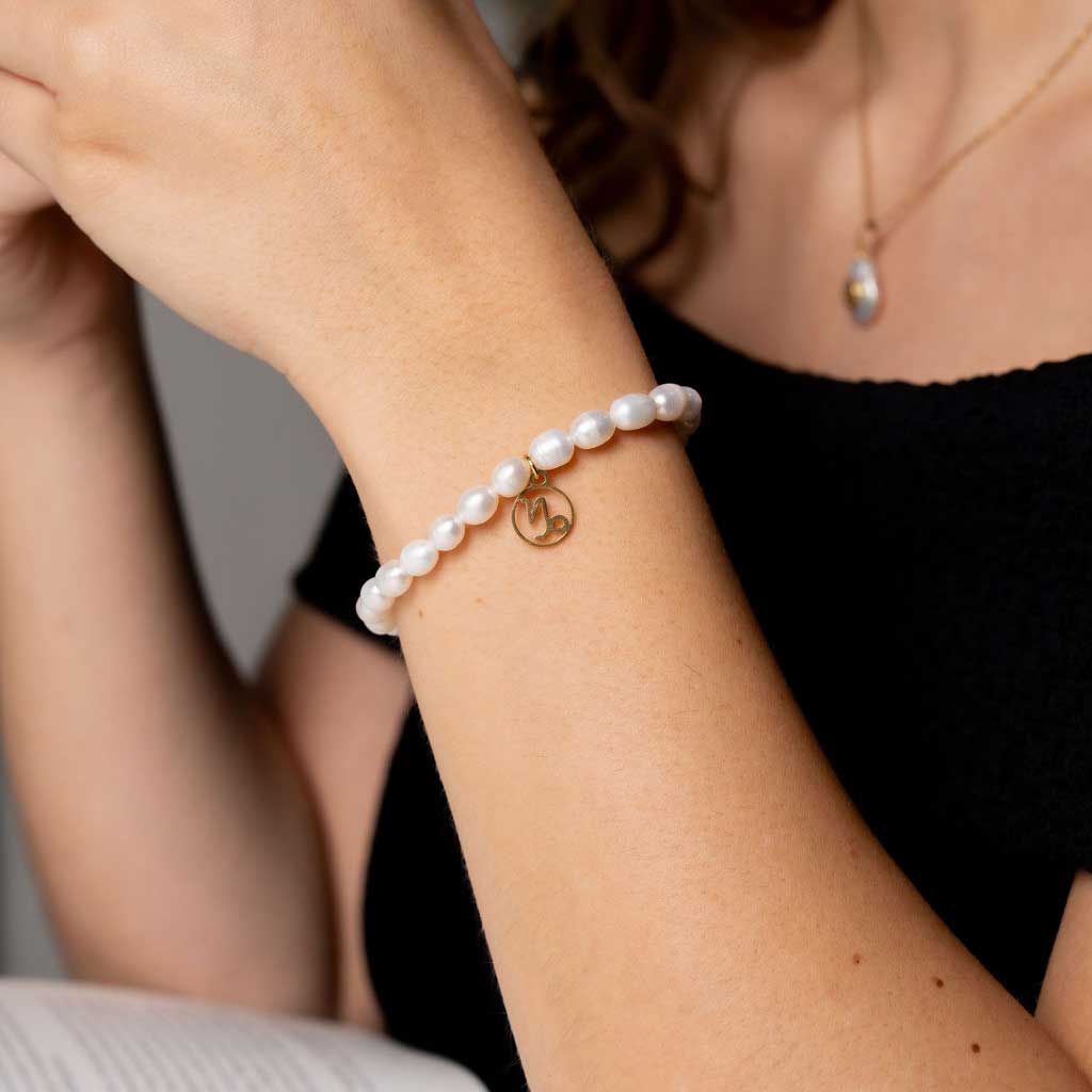 Wit parel armband met gouden edelstalen symbool sterrenbeeld steenbok op model die boek leest | Parel armband Gouden Sterrenbeeld Steenbok