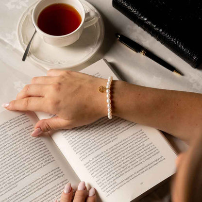 Wit zoetwater parel armband met goud edelstalen symbool van sterrenbeeld Weegschaal op model met boek | Armband Gouden Sterrenbeeld Weegschaal 