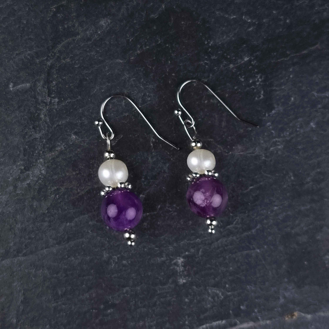 Witte zoetwater parel oorbellen met amethist en sterling zilver (925) liggend op leisteen | Amilia Amethyst