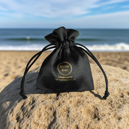 Zwart satijnen sieraden zakje met gouden logo Zhen Zhu liggend op rots met op de achtergrond de zee