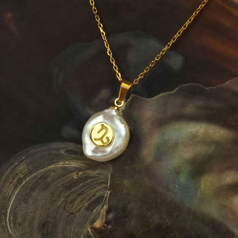 Detail van zoetwater parelketting met witte parel hanger met gouden symbool van sterrenbeeld steenbok en goud edelstaal liggend in schelp | Gouden Sterrenbeeld Steenbok