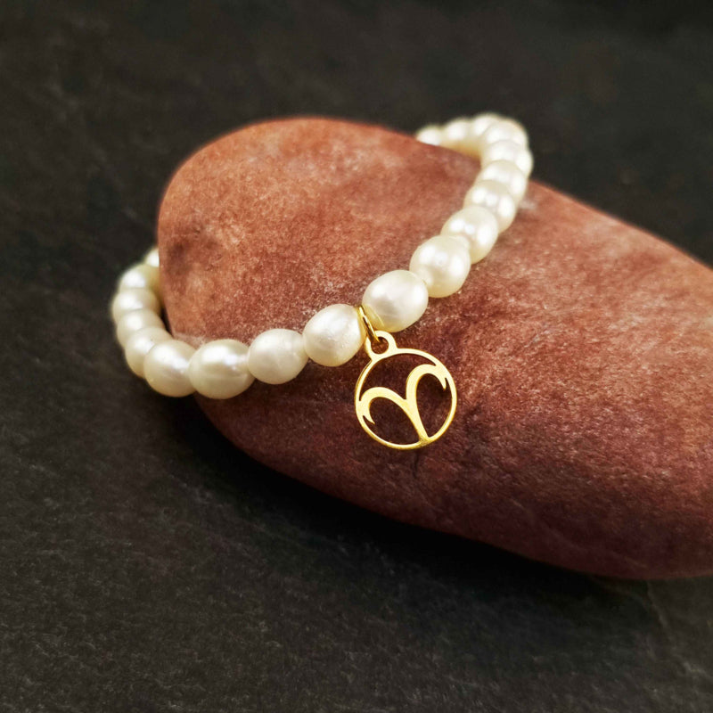 Wit zoetwater parel armband met goud edelstalen bedeltje van sterrenbeeld Ram, elastisch parel armband liggend op rode steen | Gouden sterrenbeeld Ram