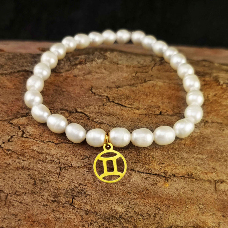 Wit zoetwater parel armband met goud edelstalen bedeltje van sterrenbeeld Tweeling liggend op hout | Gouden Sterrenbeeld Tweeling