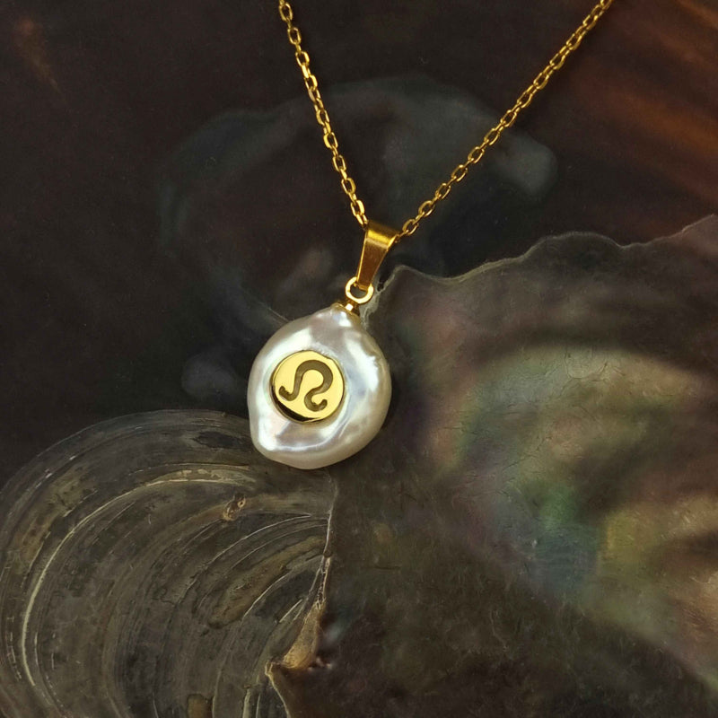Detail van zoetwater parelketting met witte parel hanger met gouden symbool van sterrenbeeld leeuw en goud edelstaal liggend in schelp | Gouden Sterrenbeeld Leeuw