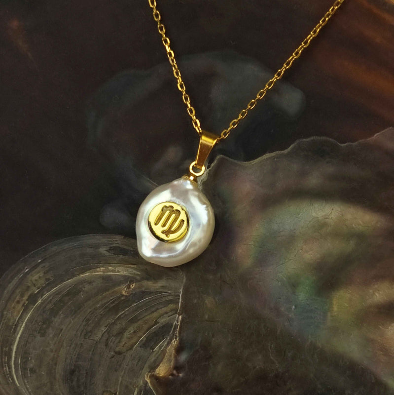 Detail van zoetwater parelketting met witte parel hanger met gouden symbool van sterrenbeeld maagd en goud edelstaal liggend in schelp | Gouden Sterrenbeeld Maagd
