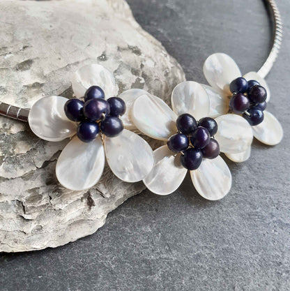 Detail van zoetwater parelketting met blauw zwarte parels en wit parelmoer in de vorm van een bloem met zilver edelstaal liggend tegen schelp | set Florien B